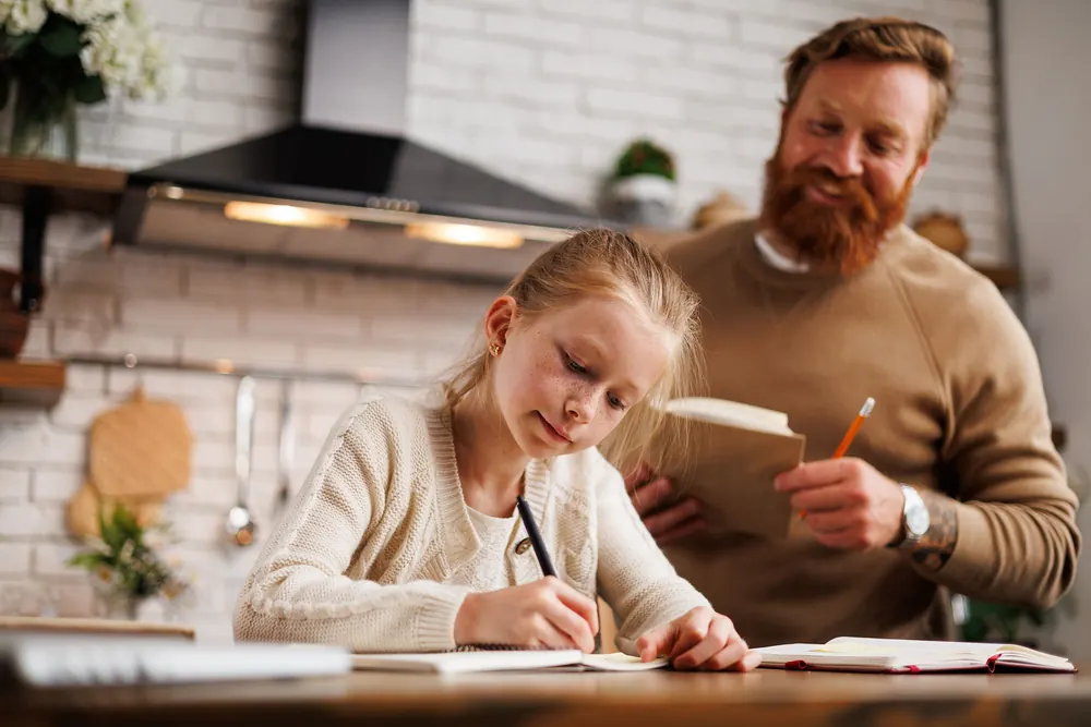 A father looks over his daughter's shoulder as she writes at the kitchen table